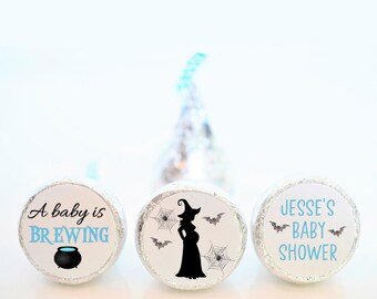 May include: Three round, silver-coloured chocolate wrappers with printed labels. The first label says "A baby is BREWING" with a black cauldron. The second label has a black silhouette of a pregnant witch with a hat and bats flying around her. The third label says "JESSE'S BABY SHOWER" with bats.