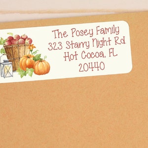 Pode incluir: Um rótulo de endereço de devolução branco com uma ilustração de outono de uma cesta de maçãs vermelhas, duas abóboras e uma lanterna. O texto no rótulo diz "The Posey Family, 323 Starry Night Rd, Hot Cocoa, FL 20440".