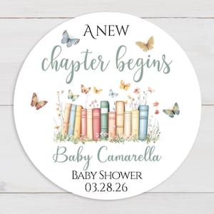May include: A round sticker with the text "A New Chapter Begins" in black and green script. The sticker features a watercolor illustration of a stack of books, butterflies, and flowers. The text "Baby Camarella" and "Baby Shower 03.28.26" are also included.