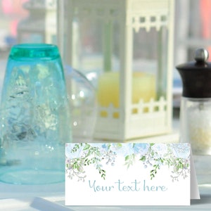 May include: A light blue place card with a watercolour floral design featuring white flowers and green leaves. The text "Your text here" is printed in a light blue font on the card.