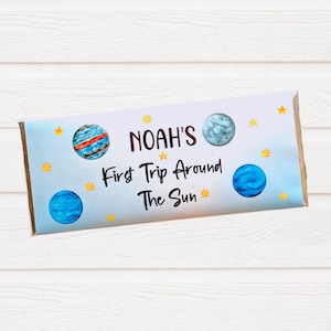 May include: A white chocolate bar wrapper with a blue background and yellow stars. The wrapper has a blue and red planet, a blue planet, and a gray planet. The text on the wrapper reads "Noah's First Trip Around The Sun".