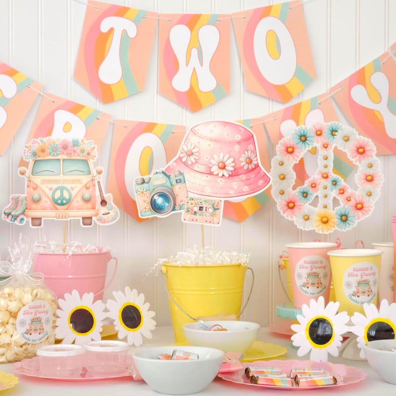May include: A colorful party scene with a banner that says "TWO". Decorations include a peace sign made of flowers, a flower-adorned hat, a camera, and a vintage van. Pink and yellow buckets, daisy cutouts, and cups complete the festive look.