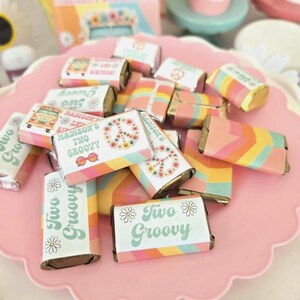 May include: A collection of wrapped chocolate bars with colorful, retro-themed packaging. The wrappers feature phrases like "Two Groovy" and "Madison's Two Groovy," along with daisy and peace sign graphics. The bars are arranged on a pink, scalloped-edge tray.
