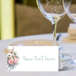 May include: A white place card with a floral design in shades of pink, white, and green. The text "Your text here" is written in light blue cursive font.