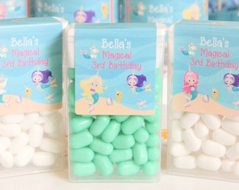 May include: Three clear plastic containers filled with pastel green and white candies. Each container has a blue and white label with the text "Bella's Magical 3rd Birthday" and a mermaid illustration.