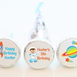 May include: Three round, silver foil-wrapped chocolate candies with colorful printed labels. The labels feature an astronaut, a rocket ship, and a planet with stars. The text on the labels reads "Happy Birthday Hunter", "Hunter's 5th Birthday", and a planet with stars.