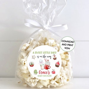 Może przedstawiać: Biała torebka na prezenty wypełniona popcorn i okrągła naklejka z napisem "A sweet little baby is on the way Grace's Baby Shower". Naklejka ma zieloną ramkę i przedstawia ilustracje zimowych przysmaków, takich jak babeczki, gorąca czekolada i bałwan.