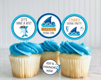 May include: Three blue and white cupcake toppers with shark designs. The toppers say "Let's Have a Bite!" "Warning Shark Zone" and "Ethan's Shark Party!"