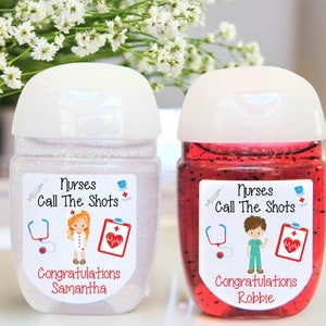 Può includere: Due bottiglie di plastica trasparente di gel disinfettante per le mani con glitter e gel rosso. Ogni bottiglia ha un'etichetta bianca con il testo "Nurses Call The Shots" e un'immagine di un cartone animato di un'infermiera. Le etichette includono anche il testo "Congratulazioni" seguito da un nome.
