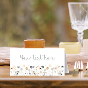 May include: White place card with the text "Your text here" in gray script font. The bottom of the card features a border of watercolor flowers in shades of peach, gray, and blue.