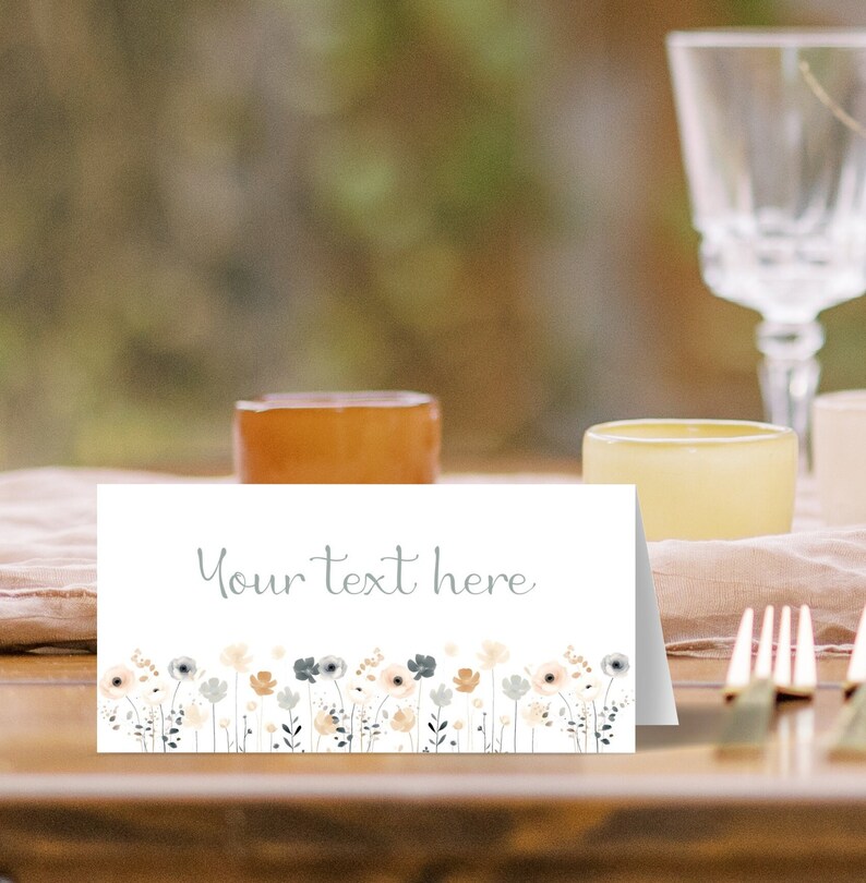 May include: White place card with the text "Your text here" in gray script font. The bottom of the card features a border of watercolor flowers in shades of peach, gray, and blue.