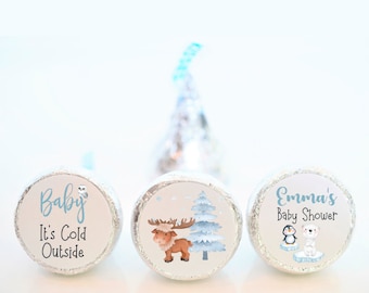 May include: Three round, silver foil-wrapped chocolate candies with printed paper wrappers. The wrappers feature winter-themed designs with a moose, a penguin and a polar bear, and the text "Baby It's Cold Outside" and "Emma's Baby Shower".