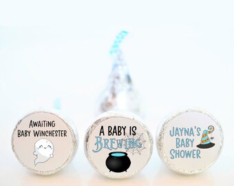 May include: Three foil-wrapped candies with custom labels. The labels feature text and graphics for a baby shower. One says "Awaiting Baby Winchester" with a ghost. Another says "A Baby is Brewing" with a cauldron. The third says "Jayna's Baby Shower".