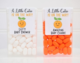 Könnte beinhalten: Zwei durchsichtige Plastikbehälter gefüllt mit orangefarbenen und weißen Bonbons. Jeder Behälter hat einen Aufkleber mit einer Cartoon-Orange und dem Text "A Little Cutie Is On The Way!" Ein Behälter hat den Text "Lily's Baby Shower" und der andere den Text "Awaiting Baby Clarke".