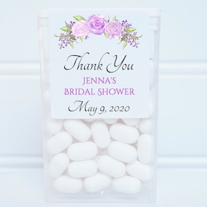 May include: A clear plastic container filled with white mints, with a label that reads "Thank You Jenna's Bridal Shower May 9, 2020". The label has a floral design with purple and pink flowers.