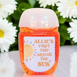 May include: A small, clear, orange-colored hand sanitizer bottle with a white label that says "Allie's First Trip Around The Sun". The label has a yellow sun with orange, blue, and green rays.