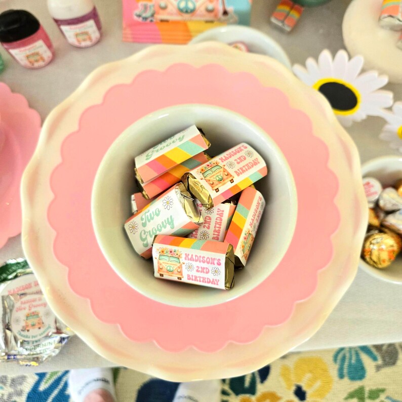 May include: A pink scalloped-edge plate with a white bowl filled with wrapped candies. The candies have colorful stripes and the text "Two Groovy" and "Madison's 2nd Birthday". Other party decorations are in the background.