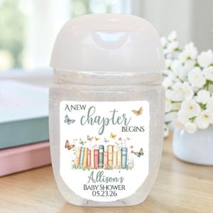 Peut inclure: Un flacon de désinfectant pour les mains transparent avec un bouchon blanc. L'étiquette indique "A New Chapter Begins" avec des illustrations de papillons et de livres. Le texte "Allison's Baby Shower 05.23.26" figure également sur l'étiquette.