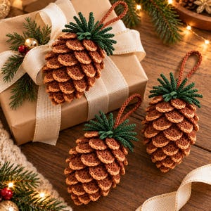 May include: Three crocheted pinecone ornaments featuring brown textured scales and dark green needle-like tops. Each ornament includes a matching brown crocheted loop for hanging, displayed alongside a gift box wrapped in tan paper.