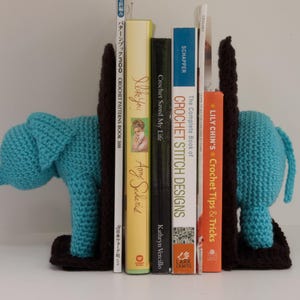 Könnte beinhalten: Ein Paar blaue gehäkelte Elefanten-Buchstützen, die einen Stapel Häkelbücher halten. Die Bücher beinhalten Titel wie "Crochet Saved My Life" und "The Complete Book of Crochet Stitch Designs".