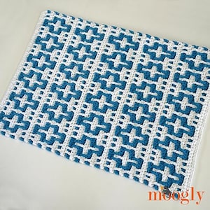 Può includere: Una coperta rettangolare all'uncinetto fatta a mano con un motivo geometrico ripetuto in bianco e blu teal. La coperta ha un bordo bianco ed è realizzata in morbido filato. Il design generale è un motivo a mosaico, che crea un aspetto strutturato.