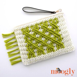 May include: A handmade crochet clutch with a white background and a green geometric design. The clutch has a black wrist strap and a gold-colored zipper. Green tassels adorn one end. The word "moogly" is in the bottom right corner.