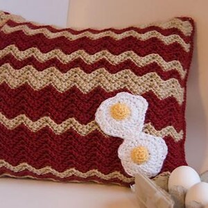 May include: A handmade crochet pillow with a red and tan chevron pattern. Two white crochet fried eggs with yellow yolks are attached to the front. The pillow is a decorative item.
