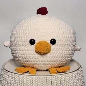Könnte beinhalten: Ein weißes, gehäkeltes Plüschtier in Form eines Huhns mit rotem Kamm, gelbem Schnabel und schwarzen Augen. Das Spielzeug sitzt auf einer weißen, gehäkelten Oberfläche.