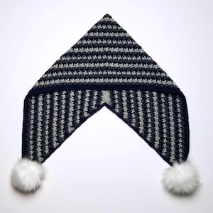 May include: A handmade crochet hat in a triangular shape, with horizontal stripes in shades of grey and navy blue. The hat has a dark navy blue border and is finished with two white faux fur pom-poms at the ends.