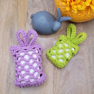 Pode incluir: Dois saboneteiros de crochê, um roxo e outro verde, com alças em forma de orelhas de coelho. Os saboneteiros são feitos com um padrão de malha e são perfeitos para segurar uma barra de sabão.