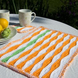 Könnte beinhalten: Ein handgefertigter, quadratischer Häkel-Tischset mit einem wellenförmigen Muster in Orange, Weiß und Grün. Zwei Zitronen, ein kleines grünes Gericht mit zwei grünen Oliven und zwei Tassen liegen auf einer weißen Tischdecke.