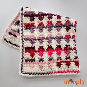 May include: A crocheted blanket with a white background and a colorful geometric pattern in shades of pink, red, and purple. The blanket has a textured, fluffy look.