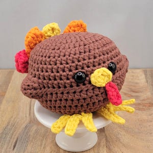 May include: A handmade crochet turkey with a brown body, orange, yellow, and red accents. The turkey has large black eyes and a yellow beak and feet. It is sitting on a small white pedestal.