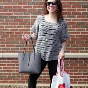 May include: A woman wearing a gray crochet top and black pants is holding a gray tote bag and a white reusable shopping bag with a red logo.