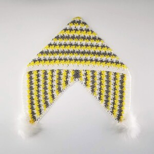 May include: A triangular crochet hood featuring alternating horizontal and vertical stripes in bright yellow, grey, and white yarn. The garment includes a solid white crocheted border along the edges and two fluffy white pom-poms attached at the bottom corners.