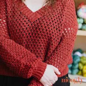 May include: A rust-colored, crocheted sweater with a V-neck design. The sweater features a textured open weave pattern and ribbed cuffs. The image shows a close-up of the sweater's details, highlighting the craftsmanship and the rich color.