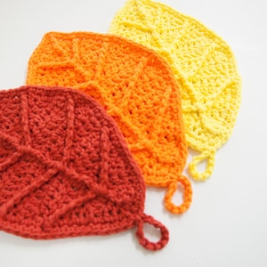 May include: Three crocheted leaf-shaped pot holders in red, orange, and yellow. Each leaf has a loop for hanging and detailed vein patterns. The pot holders are handmade and suitable for kitchen use.