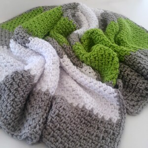 May include: A handmade crochet blanket in shades of gray, white, and green. The blanket features a textured stitch pattern and is folded to display the color blocks. The soft, chunky yarn creates a cozy and inviting look.
