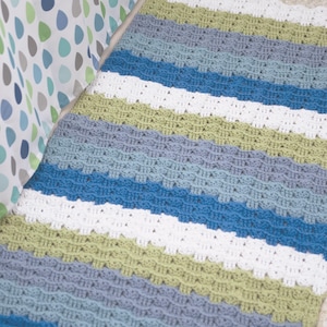 May include: A handmade crochet blanket with horizontal stripes in shades of blue, white, and green. The blanket has a scalloped edge and is likely made of soft yarn. The color palette is reminiscent of the sea and sky.