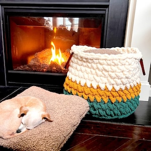 May include: A white, yellow, and green crocheted storage basket with wooden handles. The basket is sitting on a wooden floor in front of a fireplace.