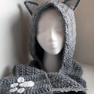 May include: A gray crocheted hood with cat ears, displayed on a mannequin head. The hood is part of a scarf, with white paw prints on the front. The hood and scarf are made of a thick, textured yarn.