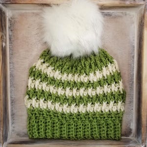 May include: A green and cream-colored crocheted beanie with a large white faux fur pom-pom on top. The hat features horizontal stripes of green and cream, and is displayed in a wooden frame.