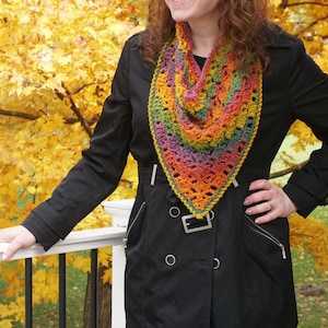 May include: A colorful, handmade crochet shawl in shades of orange, yellow, green, and purple. The shawl is triangular and worn over a black coat. The background features a vibrant display of autumn foliage.