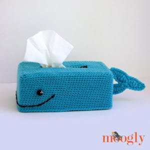 May include: A crocheted whale-shaped tissue box cover in teal blue with a black smiling mouth and black eyes. The tissue box cover has a tail fin and is sitting on a white surface.