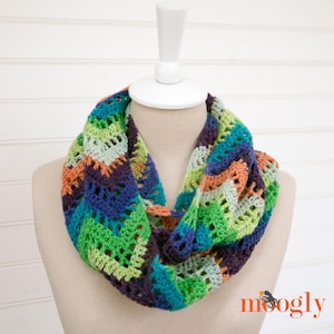 May include: A colorful crocheted infinity scarf with a multi-colored chevron pattern in shades of green, blue, purple, and orange. The scarf is displayed on a white mannequin.