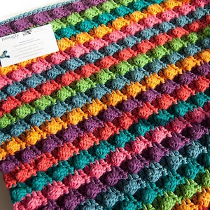 May include: A vibrant, hand-crocheted blanket with a textured bobble stitch design. The blanket displays a spectrum of colours: purple, teal, orange, pink, and green. A white tag with black text is attached to the blanket.