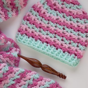 May include: A handmade crochet hat and scarf set in shades of pink and mint green. The hat is a beanie style, and the scarf is a long, rectangular shape. A wooden crochet hook is also present. The set is made of soft yarn.