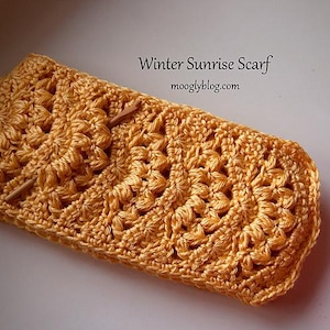 May include: A golden yellow crocheted scarf with a textured floral pattern. The scarf is secured with a wooden pin. The text "Winter Sunrise Scarf" and "mooglyblog.com" are visible on the image.