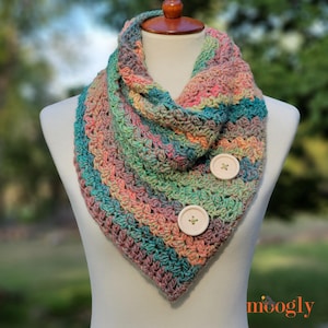 May include: A multi-colored crochet cowl scarf with button closures. The scarf features a mix of pastel colors, including pink, blue, green, and orange. Two large, round, off-white buttons secure the scarf. The word "moogly" is in the bottom right corner.