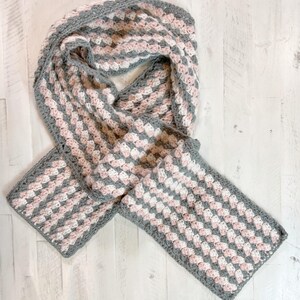 Op de afbeelding: Handgemaakte gehaakte sjaal in een granny square patroon. De sjaal heeft een herhalend patroon van roze, witte en grijze vierkanten. De randen zijn afgewerkt met een grijze rand. De sjaal is gemaakt van zacht garen en geschikt voor koud weer.